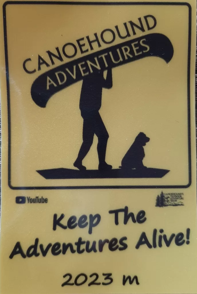 picture of a man who is camping with his dog carrying his canoe on his shoulders over a portage with the words Canoehound Adventures and Keep the Adventures Alive!