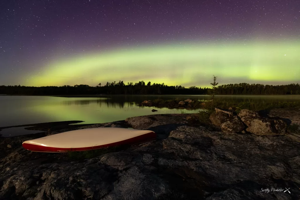 picture of a canoe on the shore of a lake while backcountry camping with a starry night sky and the northern lights