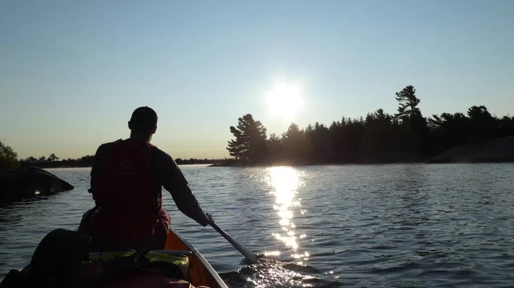 picture of a man paddling a canoe on a lake while backcountry camping as the sun is setting above some trees